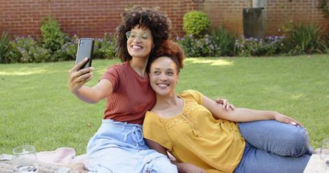 Smiling friends taking selfie while relaxing on picnic blanket in sunny backyard