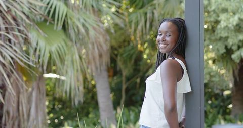 Sunlit young woman leaning on post in lush tropical garden smiling at camera, relaxed mood