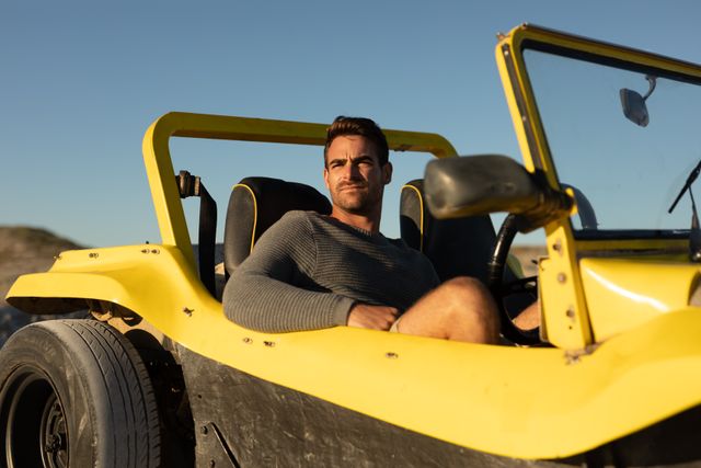 Caucasian man sitting in a yellow beach buggy on a sunny beach, admiring the view. Ideal for use in travel and adventure promotions, summer holiday advertisements, lifestyle blogs, and automotive marketing materials.