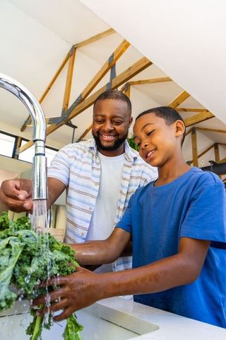 African american father and son washing kale at kitchen sink smiling and bonding