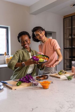 Smiling diverse friends offering colorful avocado and cabbage salad bowl in modern kitchen