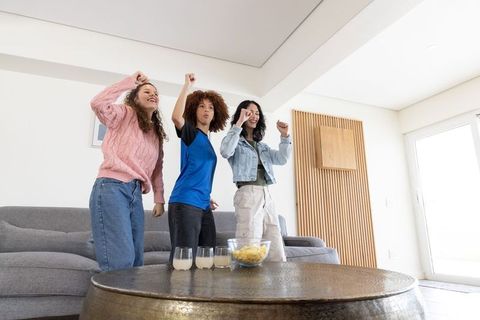 Multicultural young women celebrating at home cheering on couch with snacks and drinks