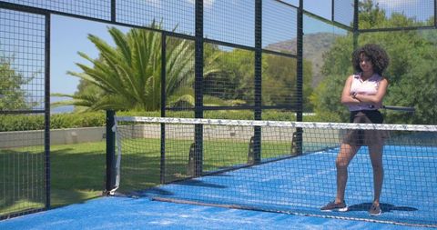 Confident woman standing with arms crossed on blue padel court holding racket, sunny sport