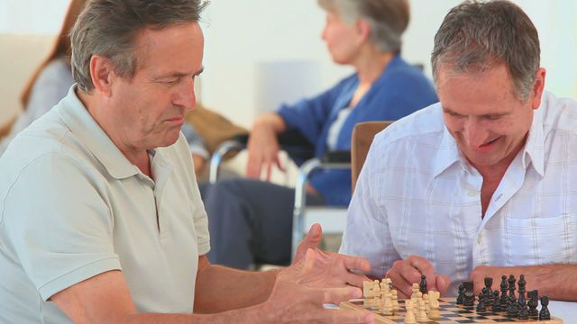 Two elderly men are playing a game of chess together indoors, engaged in a thoughtful strategy match. In the background, other senior citizens, including women in wheelchairs, are chatting and enjoying their time. This video is perfect for highlighting themes of senior living, mental engagement, hobby, friendship, and quality time in a retirement home setting.