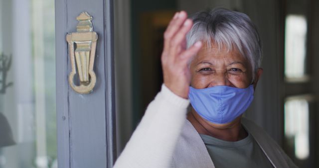 Senior woman wearing face mask greeting with a wave from her doorway. Ideal for concepts related to the pandemic, healthcare for seniors, elderly safety, and staying connected with loved ones during quarantine or isolation.