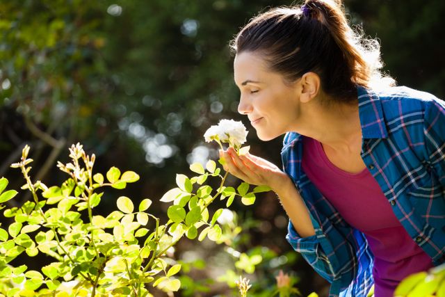 Woman enjoying the scent of roses in a backyard garden on a sunny day. Ideal for use in advertisements, gardening blogs, lifestyle magazines, and wellness content. Perfect for illustrating themes of relaxation, nature, and outdoor leisure activities.