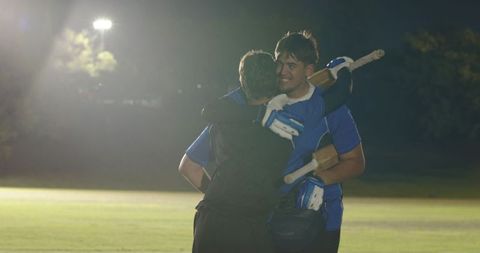 Floodlit night cricket teammates celebrating victory hugging on field holding bat and helmet