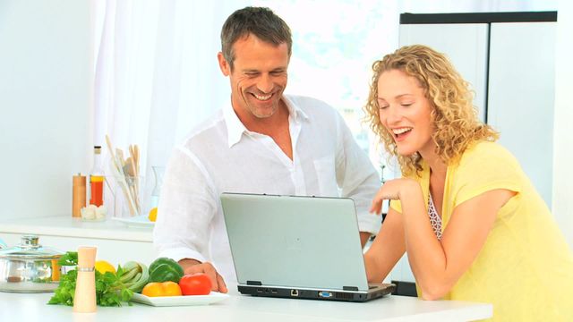 A cheerful couple is browsing recipes on a laptop in their bright, modern kitchen. Fresh vegetables are on the counter, suggesting they are planning to cook together. Ideal for advertisements on cooking websites, blogs about home cooking, technology in the kitchen, or any content centered around family life and culinary activities.