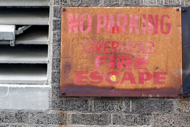Weathered 'No Parking Overhead Fire Escape' sign mounted on brick wall. Ideal for themes on urban decay, safety regulations, city infrastructure, and street scenes. Can be used in articles, presentations, or social media posts discussing urban issues, law enforcement, and public safety measures.