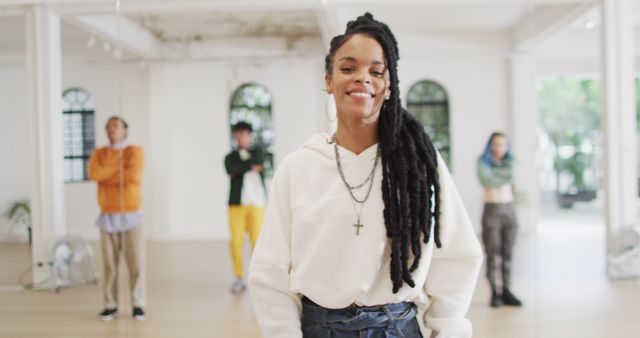 Woman with dreadlocks confidently standing and smiling in a bright, modern dance studio with diverse group in background. Used for themes like confidence, diversity, teamwork, community, dance classes.