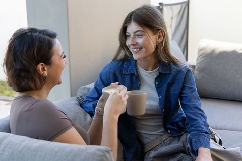 Two friends sipping coffee on cozy patio sofa, smiling and chatting