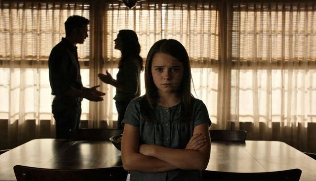 Preteen girl crossing arms at dining table while parents arguing in backlit home interior
