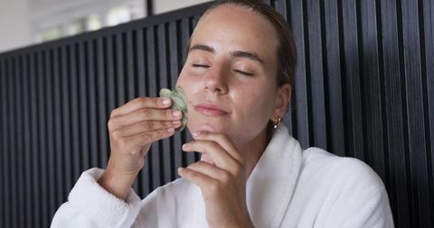 Woman applying gua sha facial massage in robe for glowing skin and selfcare routine