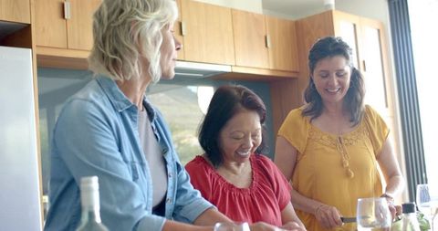 Three friends cooking together in sunlit kitchen, chopping vegetables and sharing wine