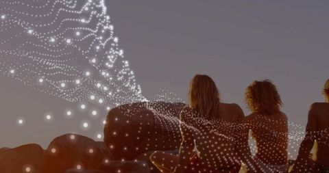 Three friends sitting on coastal boulders watching sunset with digital particle overlay