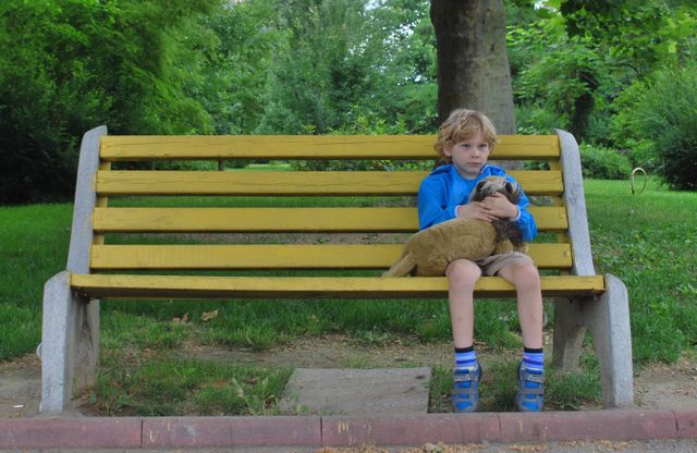 Young boy sitting on yellow park bench holding stuffed animal, surrounded by vibrant greenery. Ideal for concepts of childhood, solitude, and nature. Can be used for advertisements, articles, or blogs depicting outdoor activities, childhood moments, or emotional states related to loneliness and companionship.
