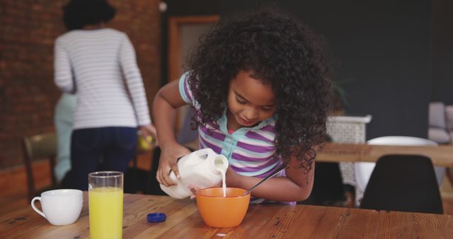 Girl with curly hair pouring milk into cereal bowl at home during breakfast. This image can be used for themes related to family routines, healthy eating, morning activities, and food advertising. Ideal for educational content on nutrition and family lifestyle.