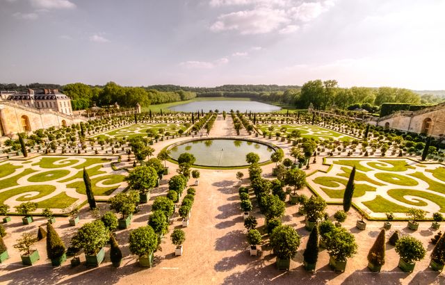 This image showcases an expansive formal garden with intricate patterns and a central fountain. The garden is adorned with trimmed bushes and tall trees, demonstrating a meticulously organized landscape. This setting is perfect for visuals related to European gardens, landscape architecture, symmetry studies, or environmental beauty. Great for use in travel guides, horticulture blogs, or urban planning presentations.