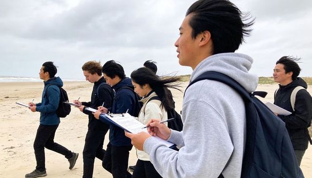 Students walking coastal fieldwork taking notes on clipboards during windy beach survey