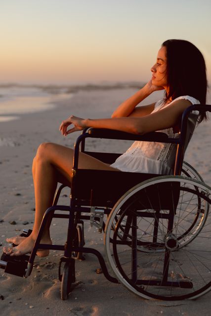Thoughtful disabled woman is enjoying a peaceful moment on the beach at sunset. This evocative scene captures serenity and contemplation as she gazes towards the ocean, offering powerful imagery perfect for promoting accessibility, independence, and inspirational themes in advertisements, articles, and social media.