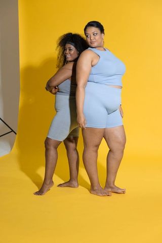 Plus-size diverse women posing back-to-back in pastel activewear on vibrant yellow backdrop
