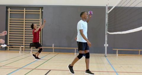 Diverse male team playing volleyball in school gymnasium serving multicolor ball
