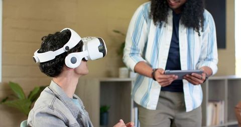 Colleague testing VR headset while coworker holding tablet in casual modern office for teamwork demo