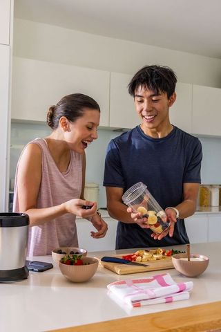 Happy asian man and african american woman preparing fruit smoothie on kitchen island