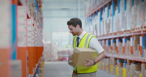 Warehouse worker scanning inventory while holding cardboard box in hi-vis vest on aisle