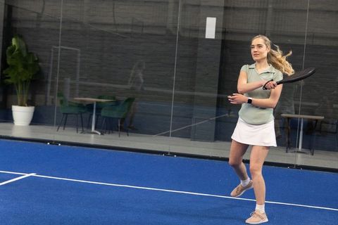 Female athlete swinging paddle on indoor blue court with glass wall and lounge reflection