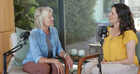 Two mature women recording podcast in bright living room with microphones and coffee