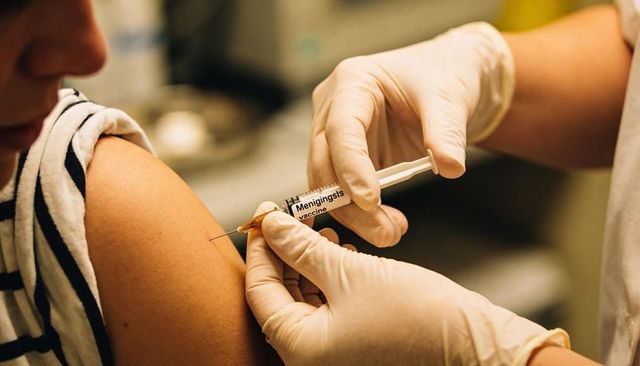 Nurse administering meningitis vaccine into upper arm of patient wearing striped top