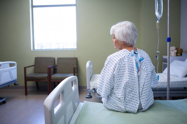 Elderly woman sitting on hospital bed, looking out window, wearing hospital gown, IV drip attached. Ideal for healthcare, medical treatment, patient care, elderly care, hospital environment, recovery process themes.