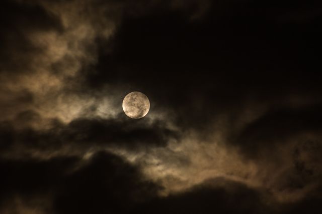 Full moon shining through thick, cloud-covered night sky creating an eerie and mysterious atmosphere. This could be used in designs for Halloween, nighttime scenes, or any content requiring a spooky or moody ambiance. Suitable for backgrounds, posters, book covers, and websites.