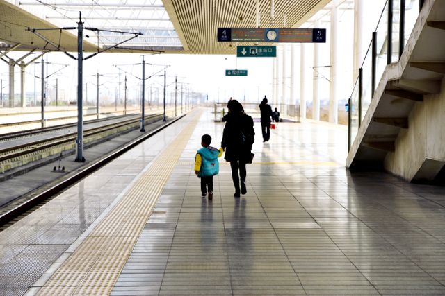 Parent and child holding hands while walking on a clean train station platform. Ideal for content about family travel, public transportation, urban commuting, and infrastructure. Could be used in a travel blog, transportation guide, or family-focused advertisement.