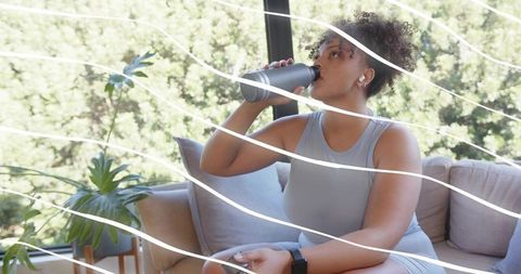 Curly-haired woman drinking from metal bottle on sofa with plants and earbuds