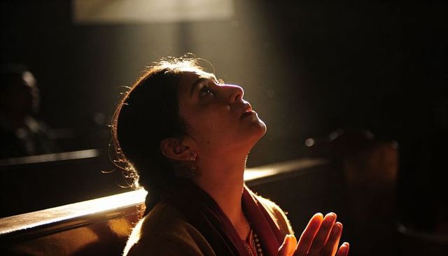 Praying indian woman sitting on wooden pew in dim chapel with sunlight beam and dust motes