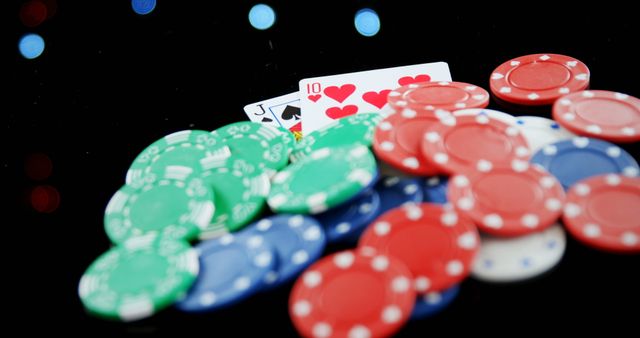 An assortment of red, green, and blue poker chips scattered on a black surface, with playing cards in the background. Useful for advertisements relating to card games, casinos, and gambling scenes. Ideal for illustrating concepts of strategy, risk-taking, and competitive gaming environments.