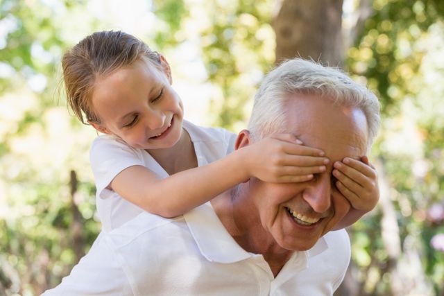 Granddaughter covering her grandfather's eyes while playing outdoors in a forest. Ideal for use in family-oriented advertisements, articles on intergenerational relationships, or promotional materials for outdoor activities and family bonding.