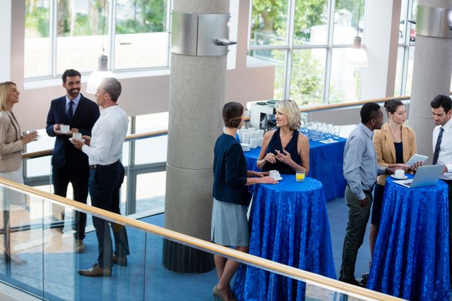 Business professionals are seen networking and interacting during a break at a conference center. The scene features people of diverse backgrounds engaged in conversation, holding coffee cups and drinks. The setting includes tables with blue tablecloths, refreshments, and a bright, modern indoor space. Ideal for themes related to corporate events, networking, teamwork, professional interactions, and business meetings. Suitable for illustrating articles, presentations, websites, and promotional materials related to business and corporate culture.