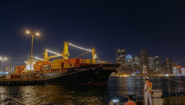 Nighttime container ship berthing at urban port with illuminated cranes and city skyline reflections
