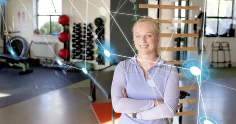 Smiling female fitness trainer crossing arms in bright gym with digital network overlay