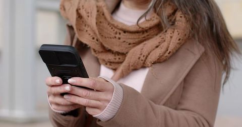 Mature woman using phone with protective case and ring grip wearing tan coat chunky scarf