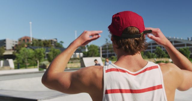 Young man wearing red cap and tank top is at a skate park enjoying a sunny day and greeting friends from a distance. This image can be used for promoting outdoor activities, teenage lifestyle, and summer fun.