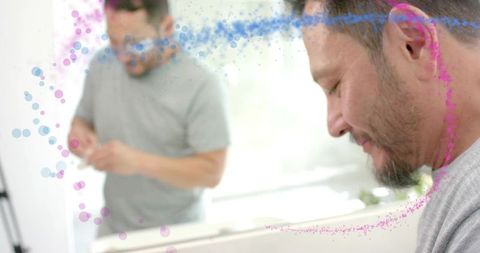 Mature man looking in bathroom mirror with friend blurred in background, colorful bokeh