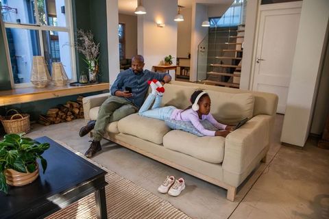 African american father relaxing with daughter using tablet and phone in modern living room
