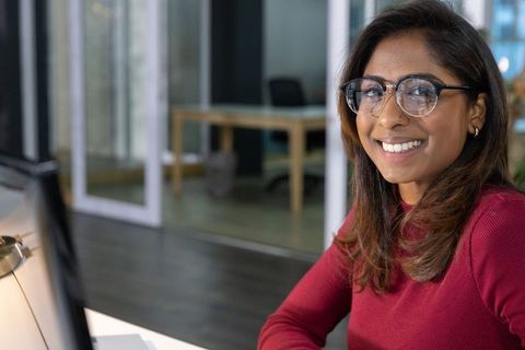 Indian professional woman smiling at camera in modern office with glasses and computer