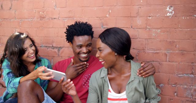 Three friends in casual clothing are laughing and enjoying each other's company while looking at a phone outdoors. They sit against a brick wall on a sunny day. This can be used in contexts promoting friendship, happiness, and social engagements. Ideal for illustrating fun moments, social media, technological interaction, or lifestyle content.