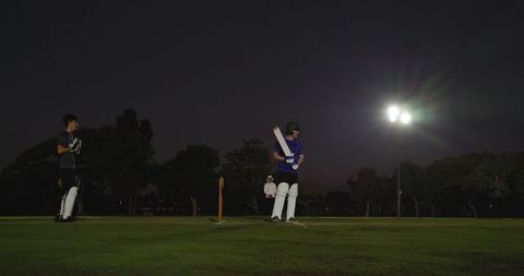 Batsman standing ready under floodlights on night cricket pitch with stumps and umpire
