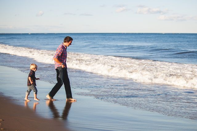 Father and son enjoying a walk along a sandy beach with the ocean waves gently crashing. Ideal for use in advertisements promoting family vacations, bonding activities, travel destinations, parenting articles, or lifestyle blogs.